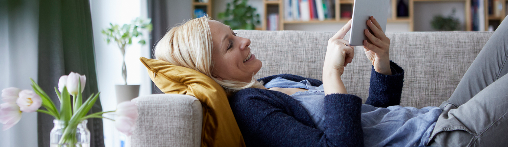 Eine Frau liegt auf einem grauen Sofa und schaut auf ihr Tablet in ihrer Hand. 