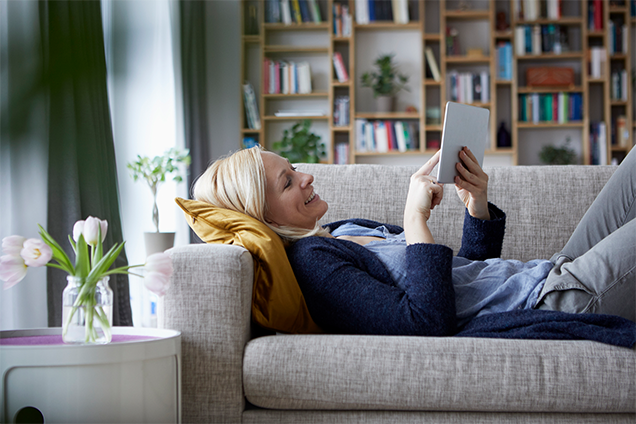 A woman is lying on a grey sofa and is looking at her tablet in her hand. 
